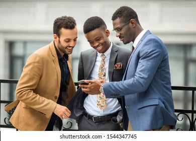 three handsome young men in suits near the building look at the phone	
