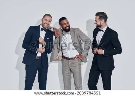 Three handsome young men in suits and bowties looking at camera and smiling while standing against gray background
