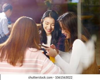 three happy beautiful young asian women sitting at table chatting talking playing with cellphone in coffee shop or tea house, shot through window glass. - Powered by Shutterstock