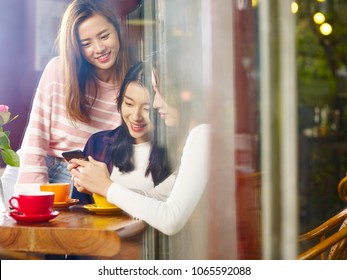 Three Happy Beautiful Young Asian Women Sitting At Table Chatting Talking Playing With Cellphone In Coffee Shop Or Tea House, Shot Through Window Glass.