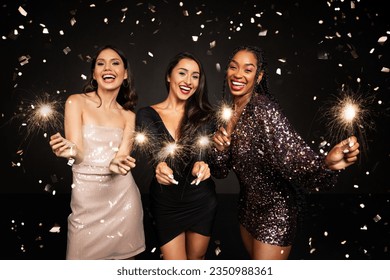 Three happy beautiful young ladies in nice dresses with bengal lights posing in confetti over black background, multiracial girlfriends celebrating New Year together, have xmas party