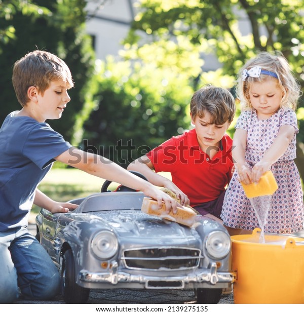 Three\
happy children washing big old toy car in summer garden, outdoors.\
Two boys and little toddler girl cleaning car with soap and water,\
having fun with splashing and playing with\
sponge.