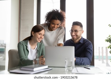 Three happy diverse colleagues working on project together, using laptop, sitting at table in office, discussing strategy, brainstorming, smiling African American businesswoman training staff