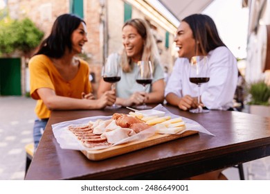 Three happy diverse women having fun together enjoying red wine at restaurant. Focus on wooden board with cheese and ham. - Powered by Shutterstock