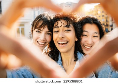 Three happy diverse young women drawing heart shape with hands outdoors. Smiling portrait of multi ethnic female friends having fun at city street. Love, youth lifestyle and friendship concept - Powered by Shutterstock