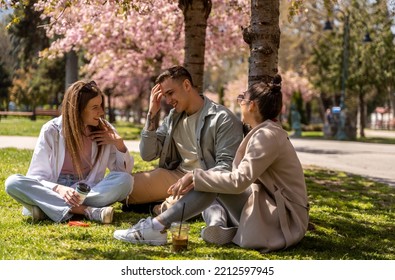 Three happy friends spending free time together in park sitting on grass and chatting