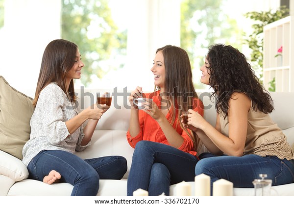 Three happy friends talking and drinking
coffee and tea sitting on a couch at
home