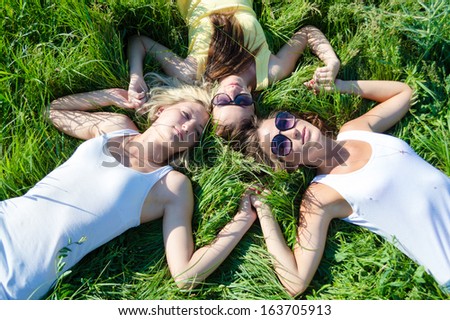 Three happy girls lying on green grass and holding hands on summer outdoors background