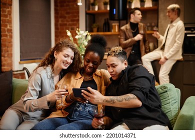 Three Happy Intercultural Girls Communicating In Video Chat At Home Party While Sitting On Couch In Living Room Against Guys Talking