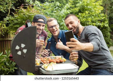 Three happy man enjoying outdoor party, standing next to barbecue grill and taking selfie. 