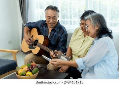 Three happy senior friends gathered indoors, one playing acoustic guitar while the others sing animatedly, female friend use smartphone photographing, enjoying music with camaraderie in living room. - Powered by Shutterstock