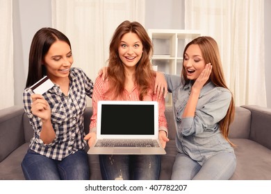 Three happy women sitting on sofa and showing laptop and bank card
