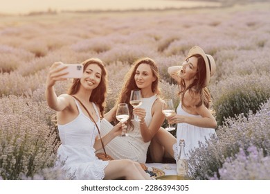 Three happy young girls with sitting at picnic and take selfie on phone on beautiful lavender field. - Powered by Shutterstock