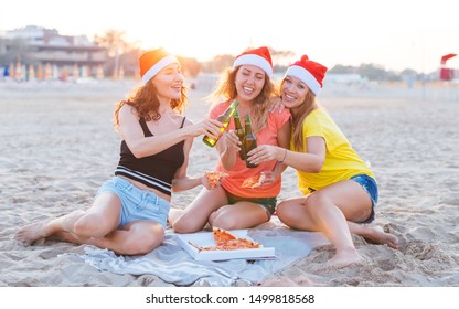 Three Happy Young Millennials Girls Celebrating Christmas On The Beach, Toasting With Beer Bottles And Eating Pizza Together, Wearing Santa's Red Hats - Xmas Under The Sun Or On Sunset Party Concept