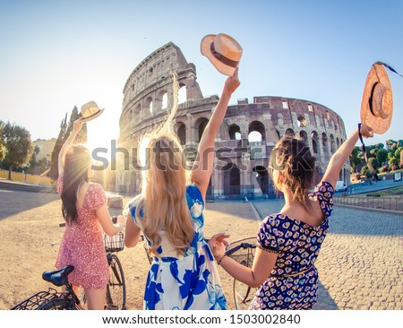 Three happy young women friends tourists with bikes waving hats at Colosseum in Rome, Italy at sunrise.