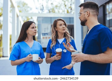 Three healthcare workers, two women and a man, in blue scrubs engage in conversation while holding coffee cups outside a medical facility. - Powered by Shutterstock