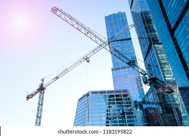 Three High-rise Cranes Against The Background Of A Glass Skyscraper Building, With An Impressive Reflection From The Sun.