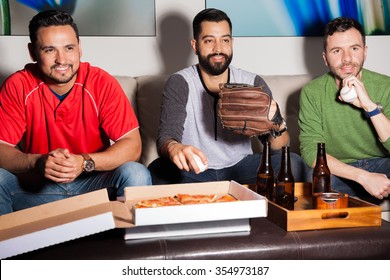 Three Hispanic Male Friends Eating Pizza And Drinking Beer While Watching A Baseball Game At Night