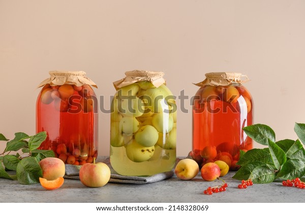Three homemade canned fruits apple and cherry\
compote in large glass jars on gray table.Summer home grown harvest\
and canned food.