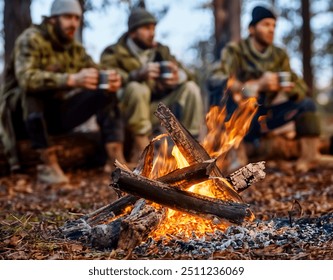 Three hunters in winter gear sit around a campfire in the forest, enjoying the warmth and hot drinks after a day of hunting - Powered by Shutterstock