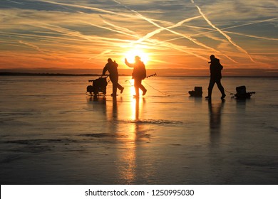 Three Ice Fishing Angler Silhouettes On Ice And Beautiful Yellow Sunset With Lines From Airplane In Pärnu Bay, Pärnu County, Estonia