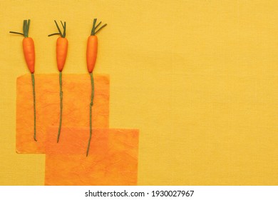 Three Imitation Carrots On A Background Of Orange Textured Translucent Paper On A Yellow Fabric Background