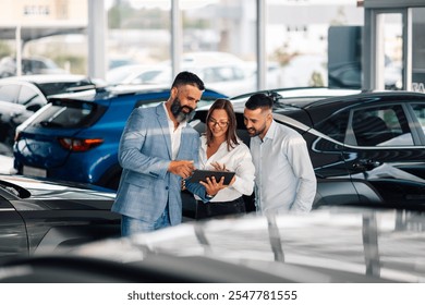 Three individuals, likely dealership staff, are collaborating using a tablet in a sunlit car showroom, amidst several new cars, displaying teamwork and modern technology. - Powered by Shutterstock