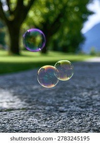 Three iridescent soap bubbles drift through the air in a park setting with green trees and a gravel path.