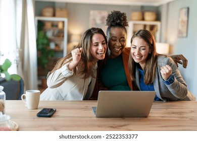 Three joyful female professionals of diverse ethnicities engage with content on a laptop. Their modern office surrounds them, reflecting a successful team effort in a corporate environment. - Powered by Shutterstock