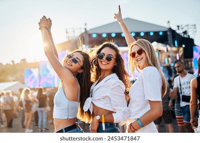 Three joyful girls dance at sunny summer beach music festival, arms raised in celebration. Female friends enjoy live concert, fun times together surrounded by crowd, ocean backdrop during sunset. - Powered by Shutterstock