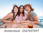 Three joyful women enjoying a sunny day together at a beachside bar, sharing laughter and friendship while sipping drinks, capturing a moment of happiness and connection in a beautiful coastline. High