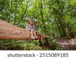 Three kids, boy and girl with caps sit atop a huge, fallen log pointing on surroundings, have fun in summer camp