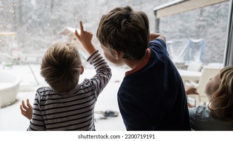 Three kids, brothers and sister, looking out the window pointing to beautiful winter nature and snowflakes falling down the sky. - Shutterstock ID 2200719191