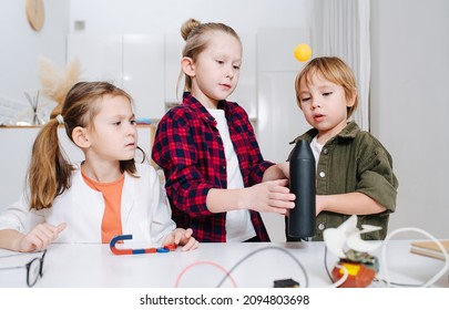 Three Kids Doing Home Science Project, Hovering Ping Pong Ball With A Fan. All Standing Behind The Table, Mezmerized With The Ball.