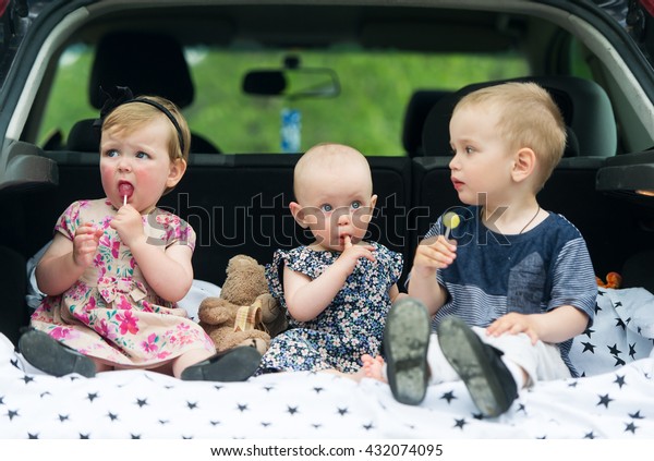 Three kids in luggage carrier of the car eat\
tasty candies.
