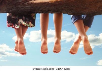 Three kids sitting in a tree dangling their feet against a blue sky in summer concept for family, friends, carefree and vacations