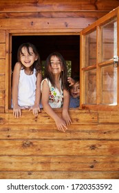 Three Kids Smiling And Playing At The Window Of A Wooden Garden House Or Tree House On A Beautiful Sunny Day.