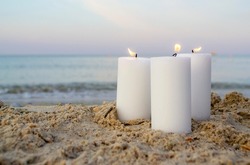 Three Large White Paraffin Candles Burn On Sand Beach Background Sea Waves. Decorative Burning Candles On Sea Coast Close Up. Candlelight, Candle Flame. Romantic Mood Backdrop. Concept Date Relaxation