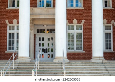 Three large white round columns at the facade entrance to a red vintage brick building. There are multiple panes of glass in tall white windows. The limestone steps have stainless steel handrails. 
