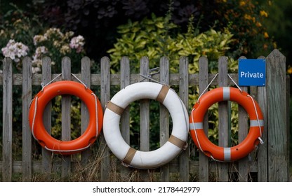 Three life buoys hang from a wooden fence with flowers and shrubs behind. Orange and white, private  - Powered by Shutterstock