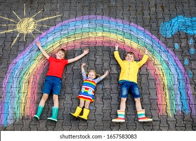 three little children  two school kids boys   toddler girl having fun and with rainbow picture drawing and colorful chalks asphalt  Siblings in rubber boots painting ground playing together 
