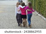 Three little girls walk hugging and talking along a pebbles path with greenbushes on the side 