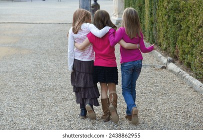 Three little girls walk hugging and talking along a pebbles path with greenbushes on the side  - Powered by Shutterstock