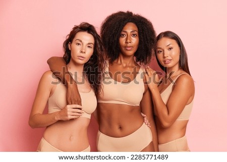 Three lovely model girls dressed in beige-colored underwear are standing embracing on a pink background, happy time friendship concept, copy space, high quality photo