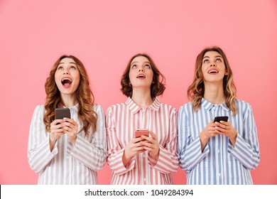 Three Lovely Young Girls 20s Wearing Colorful Striped Pyjamas Looking Upward And Using Cell Phones During Happy Sleepover Isolated Over Pink Background