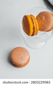Three macarons sit on a white surface, two on a glass dish.