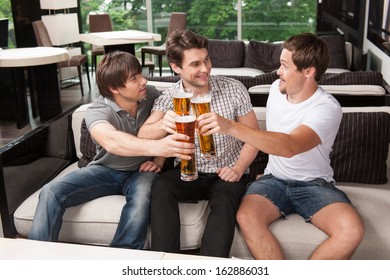 Three Male Friends Drinking Cold Beer In Hot Weather. Sitting At The Pub  