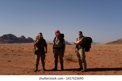 Three Male Friends Posing Together In Wadi Rum Desert. Heavy Big Backpacks, Military Clothing, Red Keffiyeh, Army Boots On Them. Sand, Rocks, Dry Grass And Sandstone Mountain Range On Background.