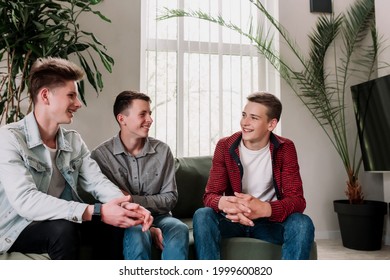 Three Male Friends Are Talking At The Green Sofa In Living Room. Guys Having A Talk, Sharing And Discussing News, Cheeling Together. Gossip Teens