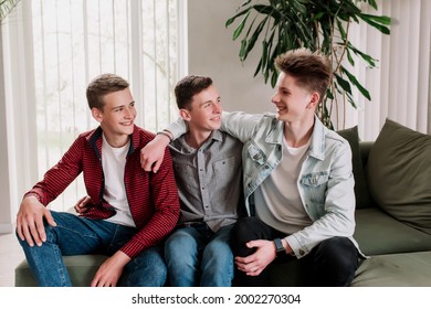 Three Male Friends Are Talking At The Green Sofa In Living Room. Guys Having A Talk, Sharing And Discussing News, Cheeling Together. Gossip Teens Hug Each Other